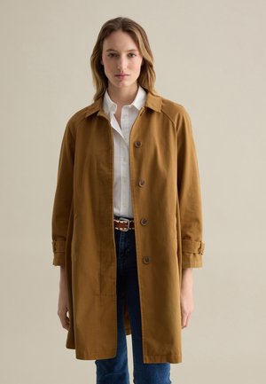 Abrigo de algodón marrón con cuello, mangas largas, cierre de botones en la parte delantera y aberturas laterales. Combinado con una camisa blanca y jeans azul.