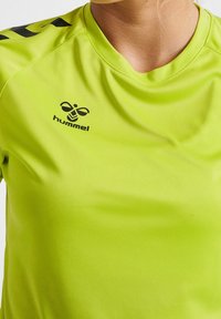 Camiseta atlética de un brillante color verde lima, hecha de una tela suave. Presenta un cuello redondeado y el logotipo negro de Hummel en la parte frontal. Acentos negros en las mangas.