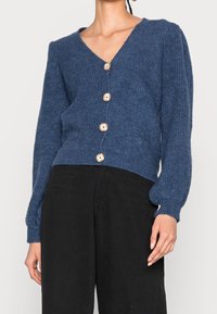 Cardigan bleu tricoté avec un col en V, cinq boutons en bois, texture côtelée et manches bouffantes, assorti à un pantalon noir.