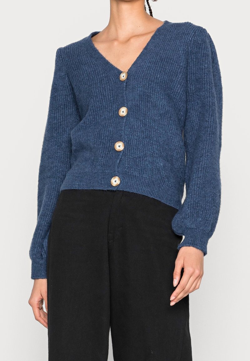 Cardigan bleu tricoté avec un col en V, cinq boutons en bois, texture côtelée et manches bouffantes, assorti à un pantalon noir.