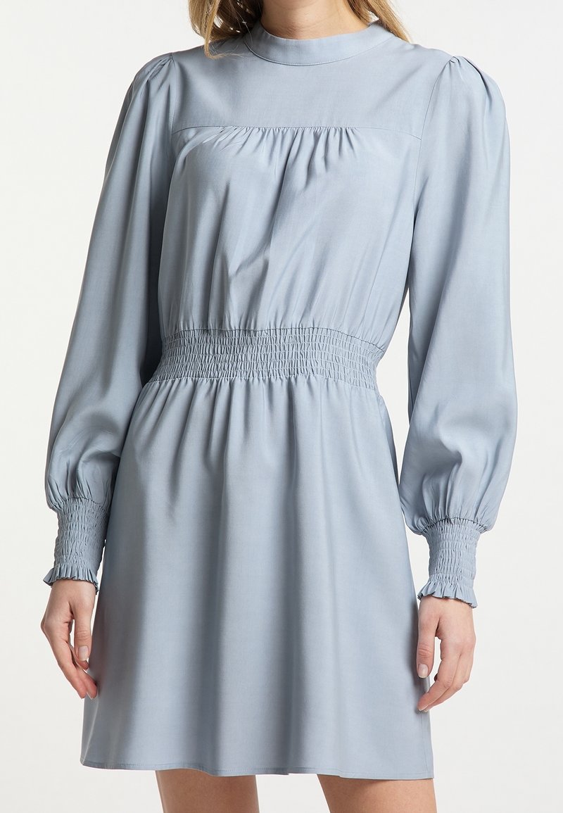 Hellblaues langärmliges Kleid mit puffärmel, hohem Kragen, geraffter Taille und gerüschten Manschetten, getragen von einer Person vor einem schlichten Hintergrund.