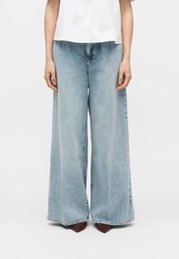 Άτομο που φοράει ανοιχτό μπλε τζιν παντελόνες με φαρδιά μπατζάκια, σετ με καφέ μυτερά παπούτσια και λευκό τοπ, στέκεται μπροστά σε μονόχρωμο φόντο.