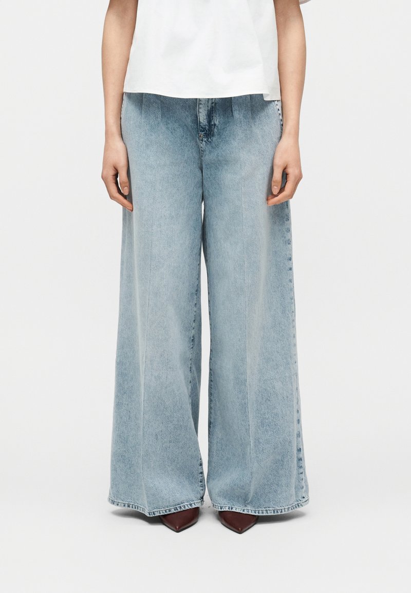 Άτομο που φοράει ανοιχτό μπλε τζιν παντελόνες με φαρδιά μπατζάκια, σετ με καφέ μυτερά παπούτσια και λευκό τοπ, στέκεται μπροστά σε μονόχρωμο φόντο.