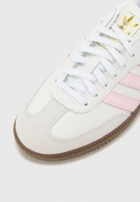 Sapatilha atlética branca com detalhes em rosa, biqueira de camurça e sola de borracha texturizada castanha. Design com atacadores e marca na lingueta.