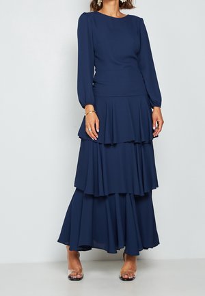 Frau trägt ein langärmliges navyblaues, gestuftes Maxikleid mit taillierter Passform und klaren, hochhackigen Sandalen vor schlichter Hintergrund.