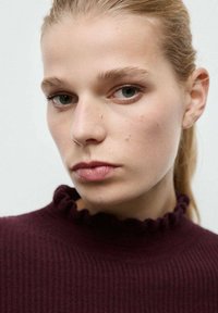 Pull en maille côtelée bordeaux avec un col volanté. Le tissu semble doux, et le modèle a un maquillage léger et naturel avec des cheveux raides.