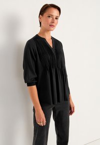 Schwarze Bluse mit Falten, V-Ausschnitt und halblangen Ärmeln mit gerafften Bündchen. Getragen mit schwarzen Hosen aus strukturiertem Stoff.