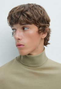 Knit turtleneck-tröja i ljusbeige, med en nära passform och ribbad hals. Modellen har rufsig brunt hår och en ren hy.