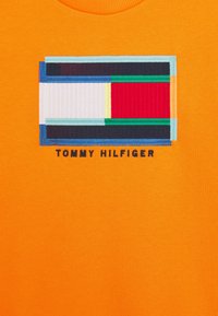 Camiseta de algodón naranja con un logotipo rectangular bordado en múltiples colores, con "TOMMY HILFIGER" en negro debajo del diseño.