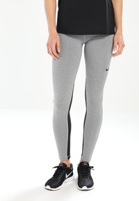 Leggings grises hechos de tela elástica con paneles laterales negros. Presenta un diseño ajustado y un logo de Nike en el muslo izquierdo superior. Un par de zapatillas negras.