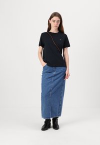 T-shirt nero a maniche corte con una spallina, abbinato a una lunga gonna in denim blu e stivali neri alla caviglia, con un dettaglio di cucitura frontale.