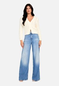 Cardigan alb tricotat, cu decolteu în V și închidere cu nasturi, asortat cu blugi largi de culoare albastru deschis, cu un model estompat.