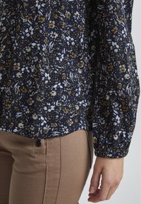 Blusa floreale blu scuro con un piccolo motivo di fiori bianchi, dorati e azzurri, maniche con polsini arricciati e orlo curvato.