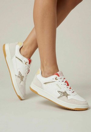 Baskets blanches basses en daim beige, détail étoile scintillante, semelle en gomme et accents rouges portées sur des jambes croisées sur un fond uni.