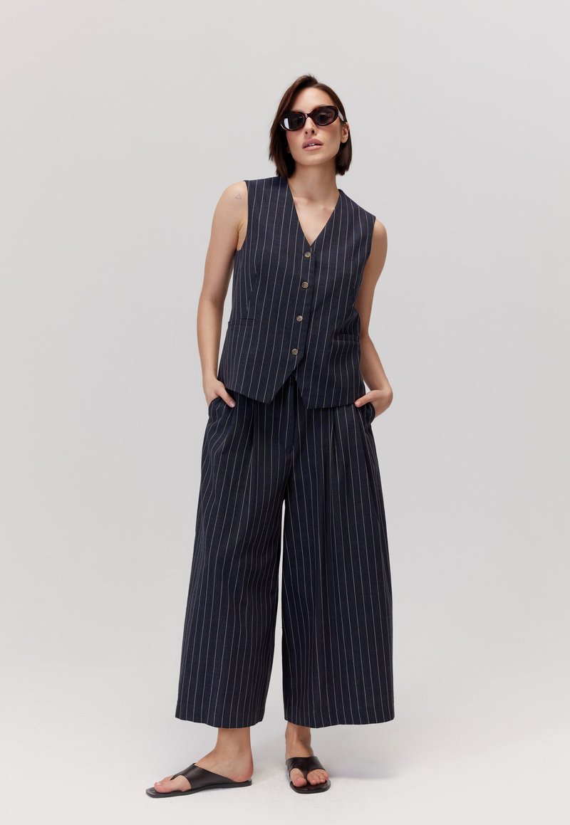 Granatowa kamizelka w paski i szerokie spodnie w lekkim materiale; guziki na kamizelce; zestawione z czarnymi klapkami i okularami przeciwsłonecznymi.