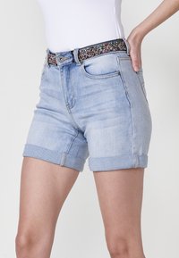Shorts en denim bleu clair avec ourlets roulés, présentant une ceinture décorative en perles et des poches avant. Design décontracté, longueur mi-cuisse.
