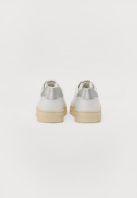 Sneaker bianche con superficie texturizzata, dettaglio argento sul tallone e suola in gomma beige. La vista posteriore mette in evidenza il design senza lacci.