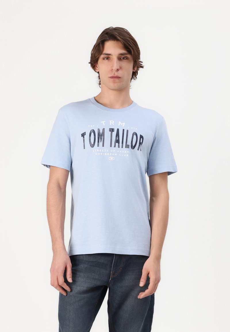 Gaiši zils kokvilnas T-krekls ar īsām piedurknēm, apaļa kakla izgriezums un priekšējā grafika melnā krāsā ar uzrakstu "TOM TAILOR" un papildu tekstu.