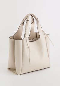 Textureret creme læder tote taske med tapered kanter, dobbelt reb-stil håndtag og hængende snore. Åben top design uden synligt hårdvarer.