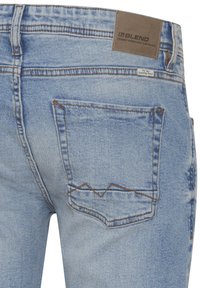 Pantalones vaqueros de mezclilla azul claro con un acabado desgastado, que presentan un bolsillo trasero con costuras angulares y una etiqueta de cuero marrón en la cintura.