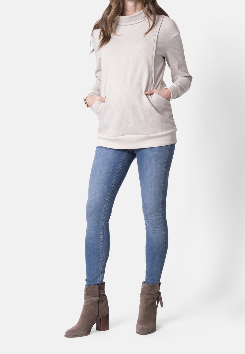 Femme portant un pull beige à col bénitier avec des poches, un jean slim bleu et des bottines en daim taupe à talons blocs.