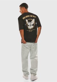 Sort oversized t-shirt med hvid due grafik på ryggen, guld tekst der står "RISE ABOVE THE REST," parret med lyseblå jeans og hvide sneakers.