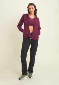 Donna che posano in una felpa maroon con zip e reggiseno sportivo, pantaloni neri e scarpe da ginnastica verdi su uno sfondo chiaro e semplice.