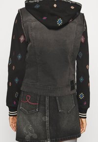 Jeansjacka med svarta ärmar, som har färgglada broderade mönster. Bakstycket är grått med ribbat avslut och svart etikett. 