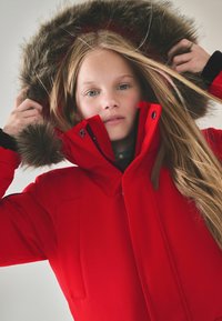 Röd parkas med pälsfodrad huva, med dragkedjestängning, framfickor och textilstruktur. Modellen håller i huvan och visar långt hår.