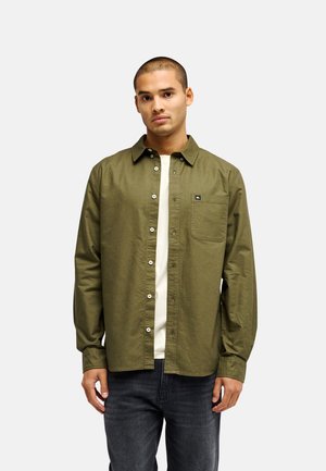 Jeune homme aux cheveux courts portant une chemise boutonnée olive ouverte sur un t-shirt blanc et un jean foncé sur un fond clair uni.