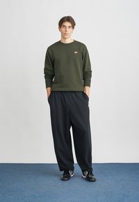 Hanorac de culoare verde măsliniu cu un mic logo roz, asociat cu pantaloni negri largi. Pantofi sport negri completează ținuta. Design simplu și casual.