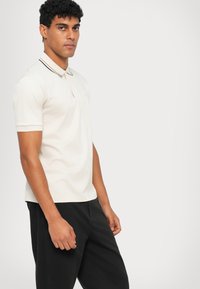 Polo-shirt in lichtbeige, met korte mouwen en een kraag met donkere strepen. Glad katoenen stof, relaxed fit, gecombineerd met zwarte broek.