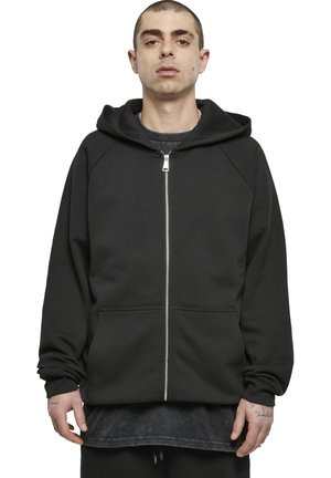 Jonge man staand met een zwarte hoodie met rits over een donker shirt, handen ontspannen langs zijn zij tegen een witte achtergrond.
