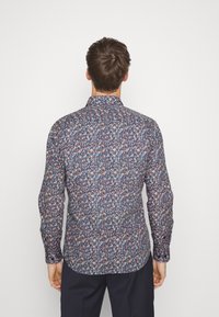 Man die met zijn rug naar de camera staat en een overhemd met lange mouwen draagt, bedekt met een dicht blauw, wit en oranje bloemenpatroon, en donkere broek.