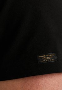 Crni pamučni majica s glatkom teksturom, sa pravokutnom etiketom zlatnim natpisom "NEOGRANIČENA SLOBODA" i ostalim detaljima.
