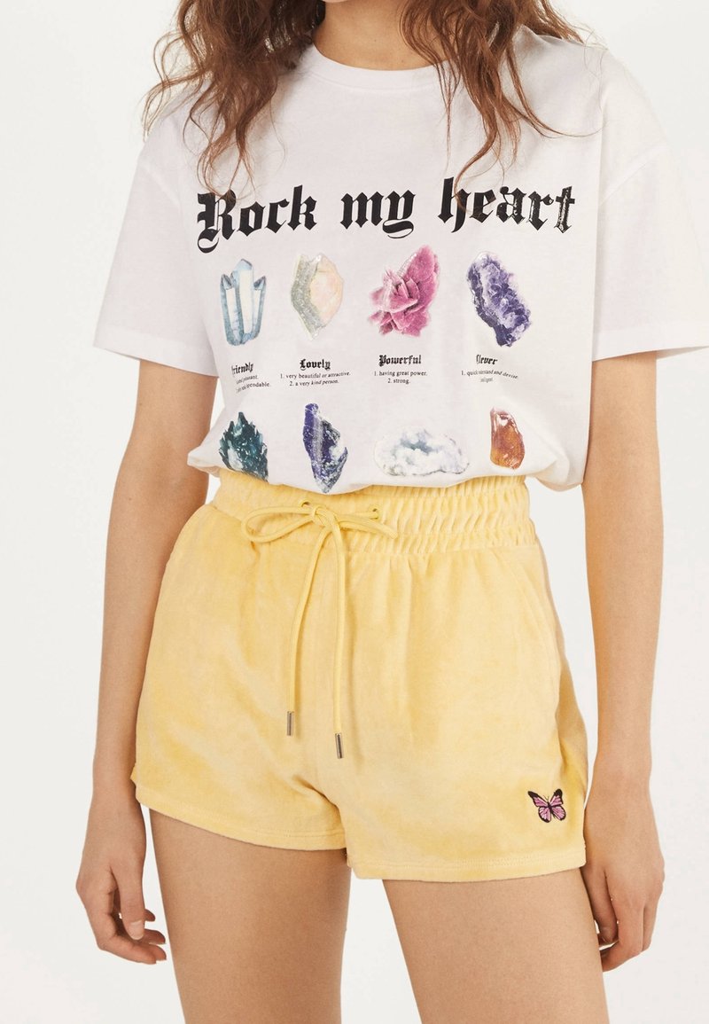 Vit t-shirt med grafisk tryck av färgglada kristaller och texten "Rock my heart". Gula shorts med dragsko och en liten fjärilsbrodering.