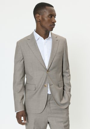 Muž v béžovom obleku a bielej košeli, stojaci s jednou rukou v vrecku, hľadí do strán na jednoliatom svetlom pozadí.