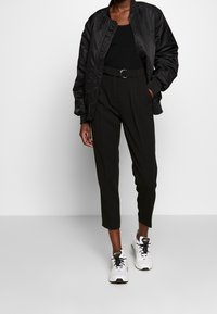 Musta bomber-takki ribbaamistaskuilla, tiukka musta neulepaita, korkeavetsiset mustat housut ja valkoiset kengät mustilla yksityiskohdilla.