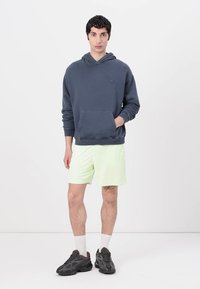 Felpa grigia scura con tasca frontale, abbinata a pantaloni corti verde chiaro. Il modello indossa sneakers nere e calzini bianchi. Design minimalista, texture liscia.