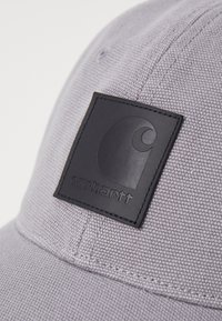 Grauerm Hut aus strukturiertem Stoff mit einem schwarzen Lederpatch, das ein Logo und den Markennamen zeigt. Auf dem Schild sind Nähte sichtbar.