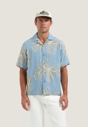 Uomo che indossa una camicia azzurro chiaro a maniche corte con grandi motivi floreali bianchi, pantaloni bianchi e un berretto da baseball beige.