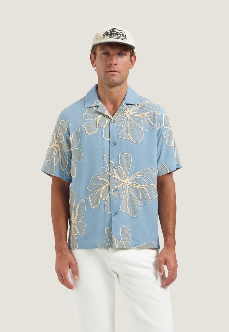 Homme portant une chemise bleu clair à manches courtes avec de grands motifs floraux blancs, un pantalon blanc et une casquette de baseball beige.