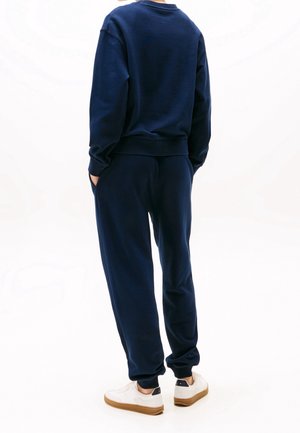 Personne portant un sweat-shirt bleu marine et un pantalon de jogging assorti avec des baskets blanches, debout les mains dans les poches, de dos.