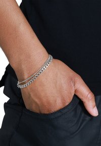 Braccialetti in argento a catena sovrapposti su un polso, con diversi design dei link. La mano è appoggiata in una tasca, su uno sfondo scuro.