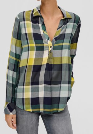 Femme portant une chemise à carreaux à manches longues verte, jaune, marine et blanche, associée à un jean gris, debout devant un fond gris clair.