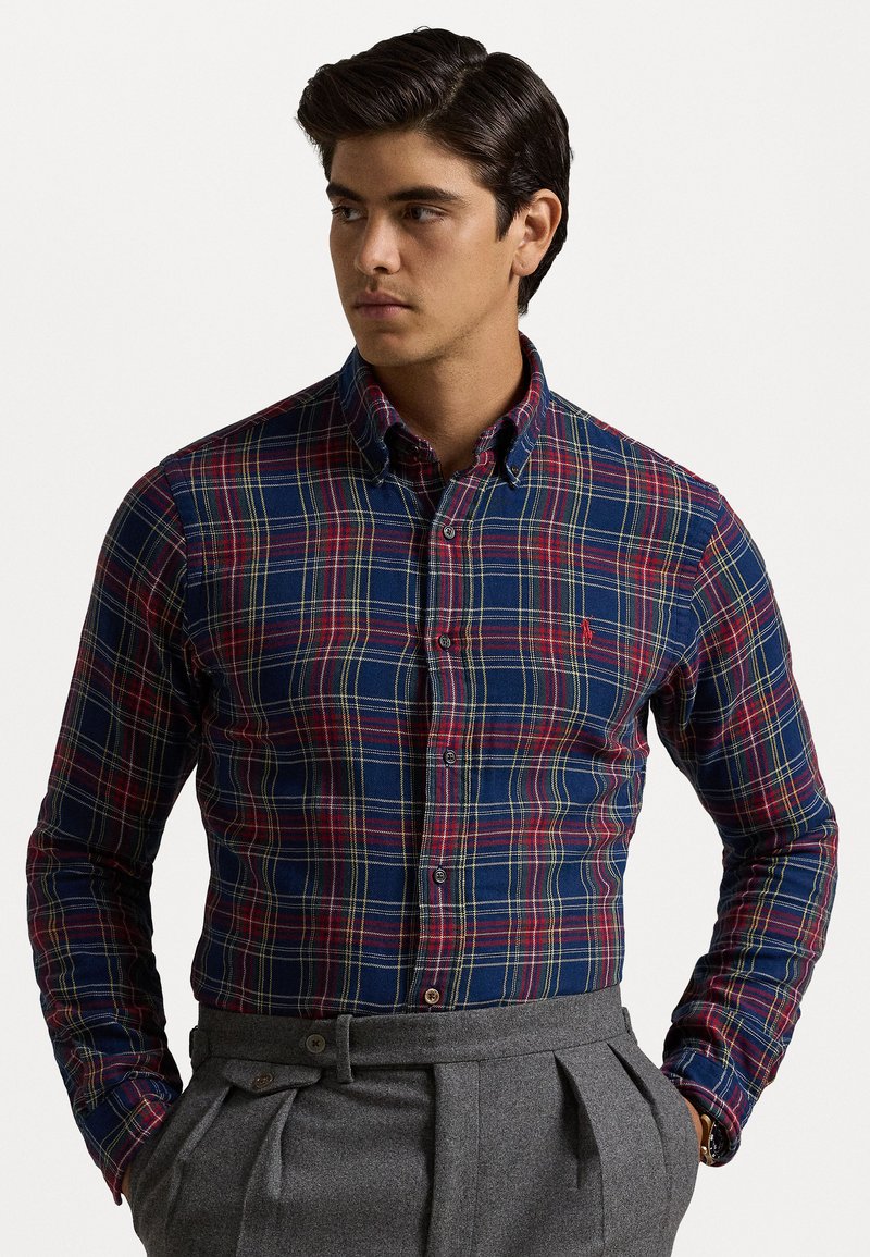 Chemise à carreaux avec motifs bleu marine, rouge et jaune. Manches longues, col et design ajusté. Associée avec un pantalon gris sur mesure.