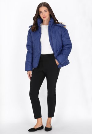 Kobieta w niebieskiej puchowej kurtce na białej koszuli, czarnych dopasowanych spodniach i czarnych balerinach, stojąca z jedną ręką w kieszeni.