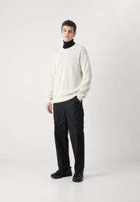 Maglione bianco a coste sopra un dolcevita nero, abbinato a pantaloni cargo neri. L'outfit presenta una vestibilità comoda e tasche pratiche.