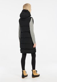 Schwarze gepolsterte Weste mit Kapuze, die ein schlankes Design und gesteppten Textur aufweist. Getragen über einem grauen Langarmshirt, kombiniert mit schwarzen Leggings und braunen Stiefeln.