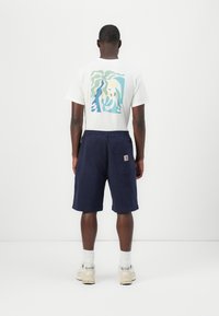 Camiseta blanca con un gráfico abstracto colorido en la parte posterior, combinada con pantalones cortos azul marino. Los zapatos son beige con detalles en gris.